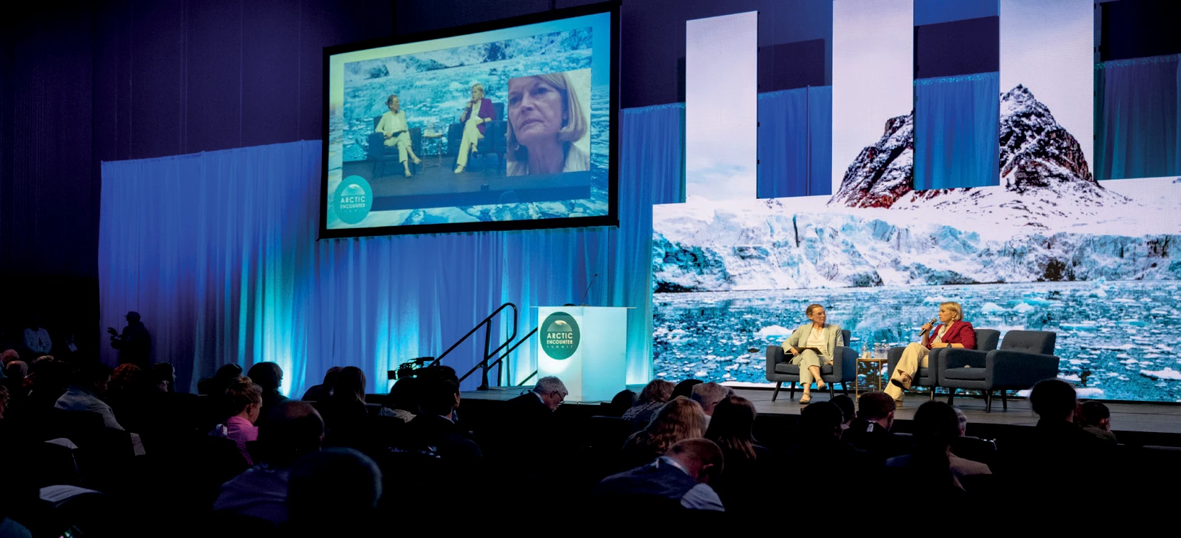 Audience attends a keynote-style conversation at the Arctic Encounter Summit, where two speakers sit on stage in armchairs with a large screen and Arctic ice landscape backdrop behind them.