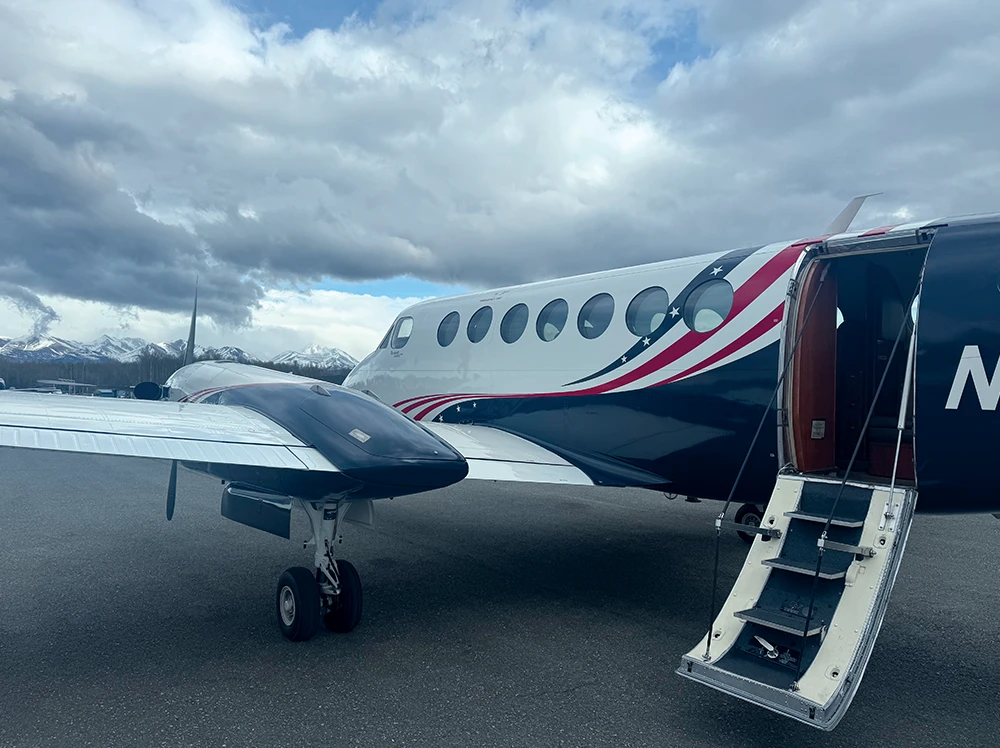 A Guardian Flight King Air 350 turboprop aircraft, featuring a blue and white fuselage with red and white star-spangled stripes, sits on an airfield with its cabin door open and airstair deployed. Snow-capped mountains are visible under a cloudy sky.
