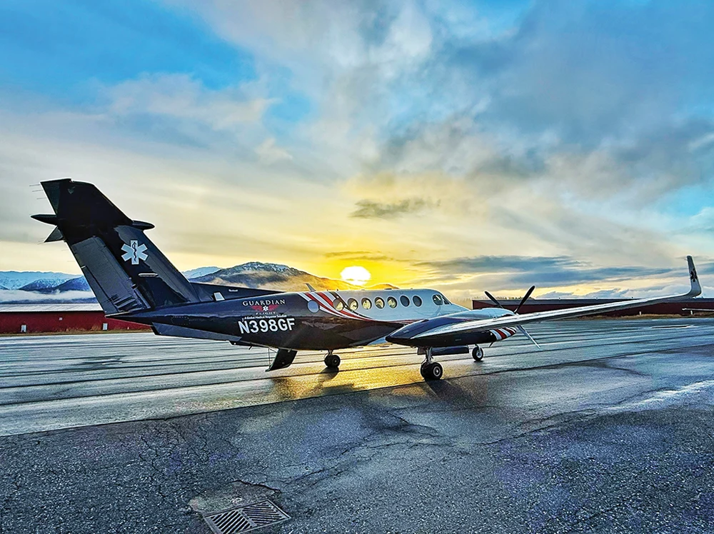 A Guardian Flight King Air turboprop aircraft (N398GF) parked on an airfield at sunset, featuring a dark blue and white fuselage with a medical Star of Life symbol on the tail. Mountains are visible in the background under a golden sky.