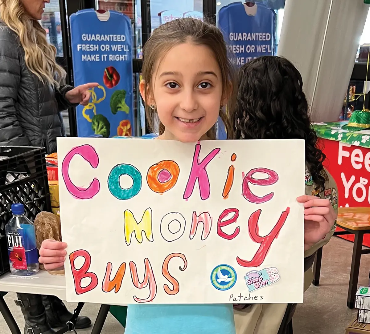 A smiling young girl holds a large, white sign with colorful, hand-drawn letters that read "Cookie Money Buys Patches" at a cookie booth inside a storefront.