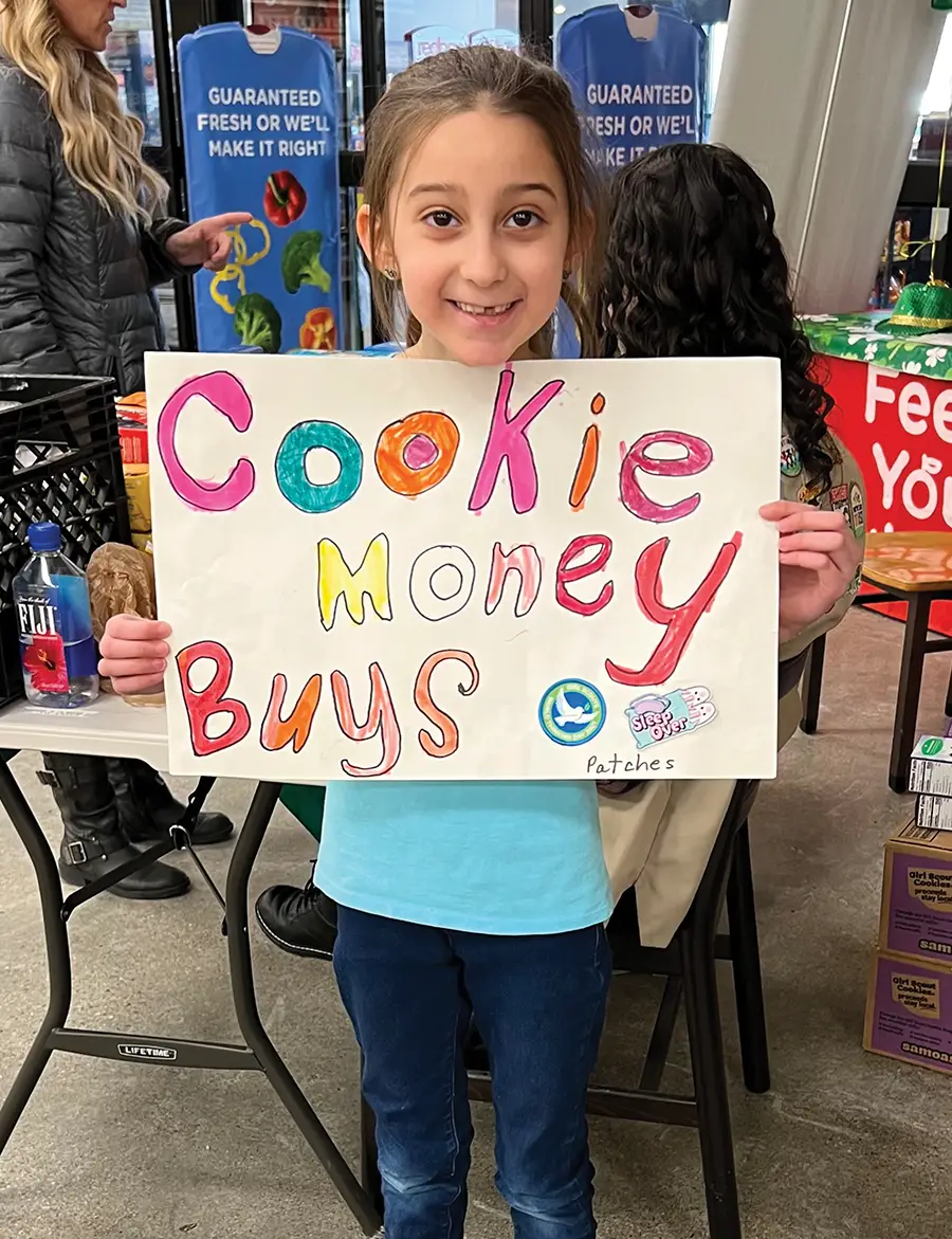 A smiling young girl holds a large, white sign with colorful, hand-drawn letters that read "Cookie Money Buys Patches" at a cookie booth inside a storefront.
