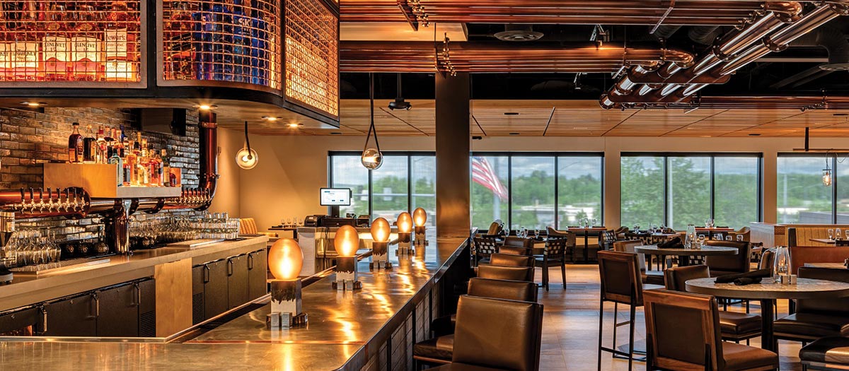Modern interior of Crimson restaurant in The Wildbirch Hotel featuring a long, polished bar with glowing lamps, leather barstools, and dining tables. Exposed copper pipes run across the ceiling. Large windows in the background overlook trees and an American flag.