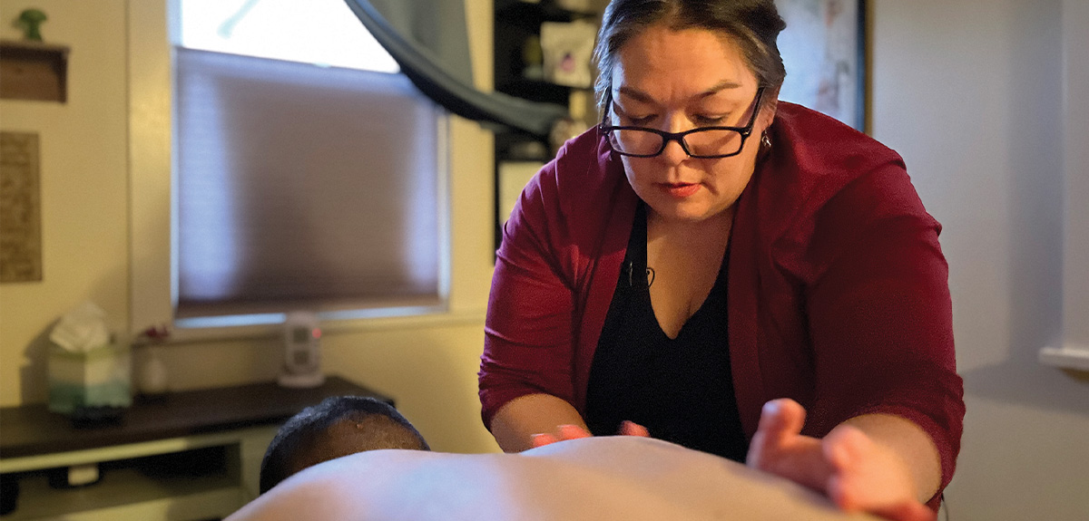 Crystal Garrett of Arctic Dawn Massage & Wellness, wearing glasses and a maroon top, performs a massage on a client's back in a dimly lit treatment room.