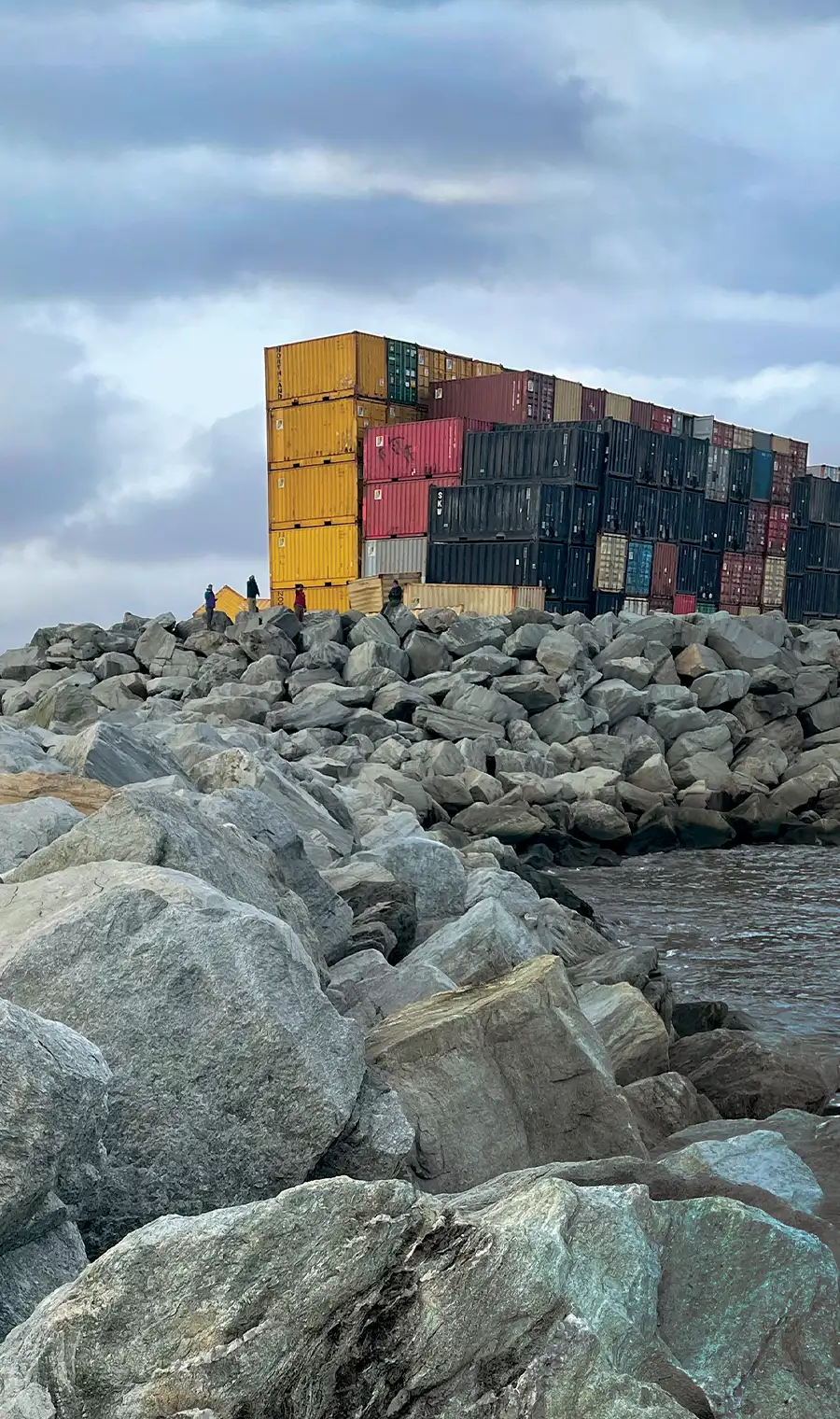 A stack of colorful shipping containers, primarily yellow, red, and black, sits atop a large rock jetty at the Port of Nome under a cloudy sky. Small figures of people stand on the rocks near the containers.