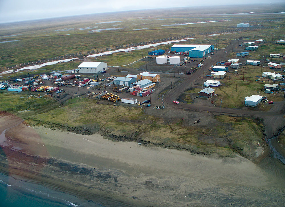 Aerial view of the coastal community of Wainwright, Alaska, showing clusters of houses, industrial buildings, and fuel tanks along a sandy shoreline and green tundra.