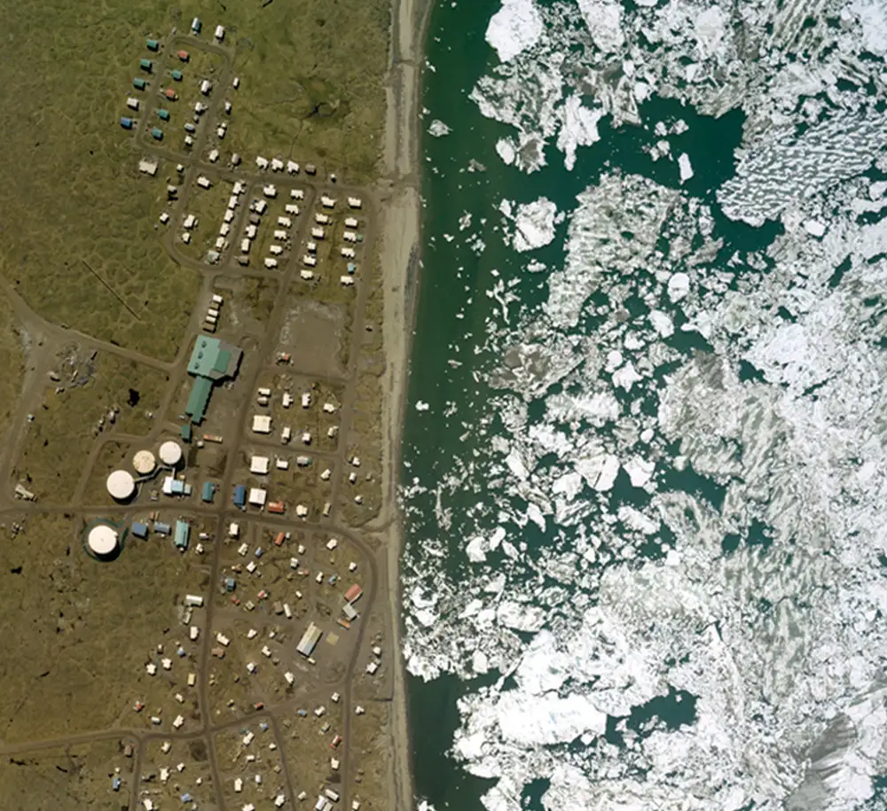 Vertical aerial view of Wainwright, showing clusters of buildings and large white storage tanks on green tundra, bordered by a sandy shoreline and the Chukchi Sea filled with white ice floes.