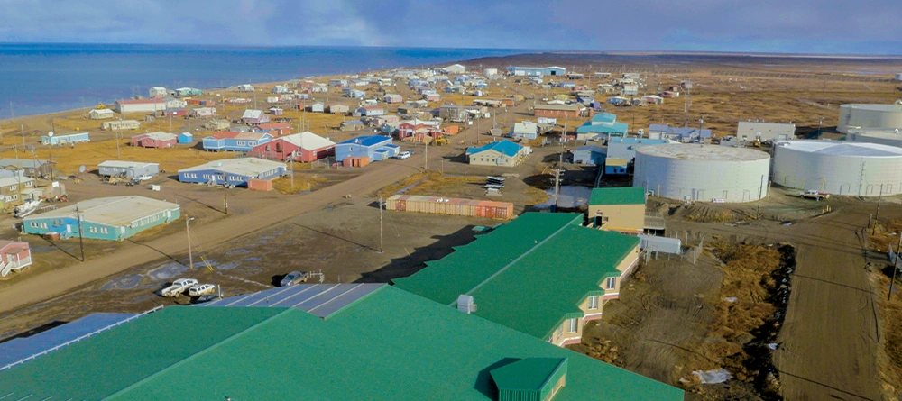 Elevated view of Wainwright, Alaska, featuring a large green-roofed building in the foreground, several white fuel storage tanks, and colorful houses along dirt roads near the coast.