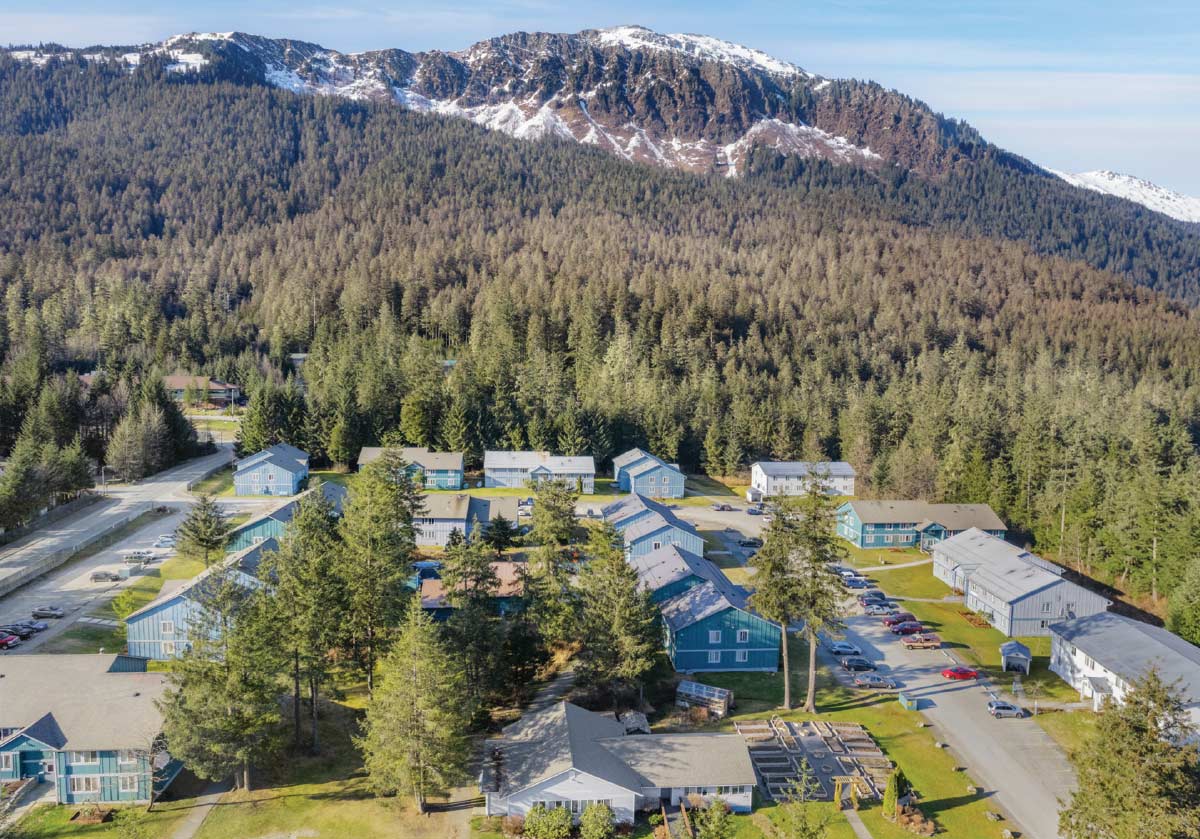 Elevated view of a multi-building apartment complex surrounded by thick pine forests, with a prominent rugged, snow-dusted mountain peak in the background.