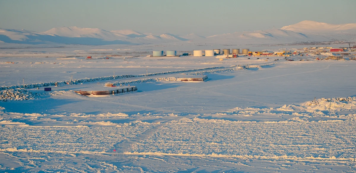 A wide panoramic view of the Port of Nome in Alaska during winter, featuring snow-covered docks, industrial storage tanks, and a background of rolling white mountains under a clear sky