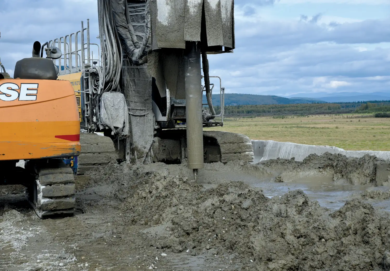 Close-up of heavy construction machinery drilling into muddy ground at an outdoor site under a cloudy sky.