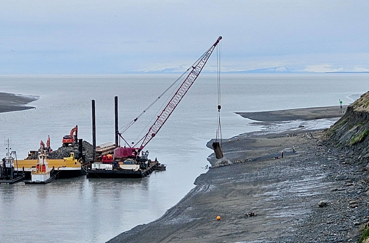 A large crane on a barge maneuvers materials along a rocky shoreline, with mountains visible across the water in the distance.