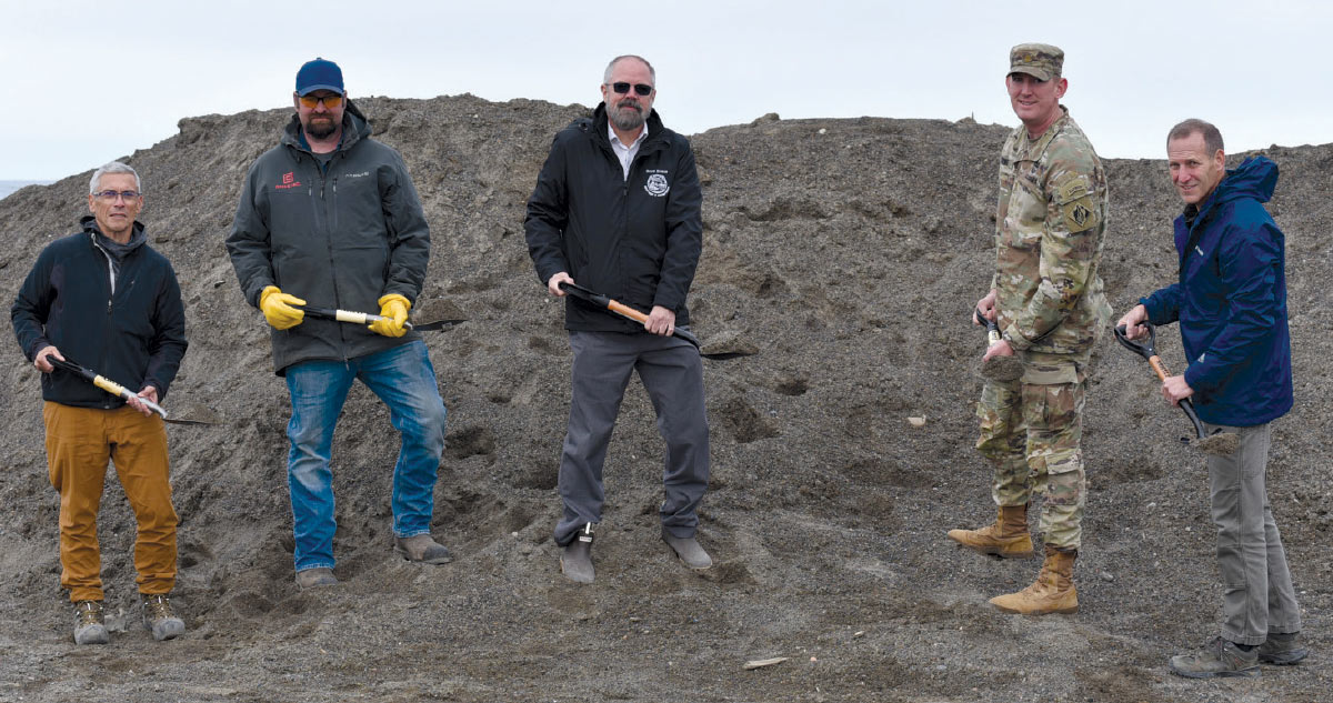 Five men, including one in military uniform, stand together holding shovels at a dirt construction site for a groundbreaking ceremony.