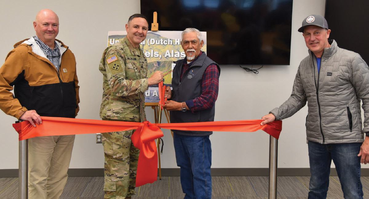 Four men, including a military officer, hold an orange ribbon and large scissors during an indoor ceremonial ribbon-cutting event.