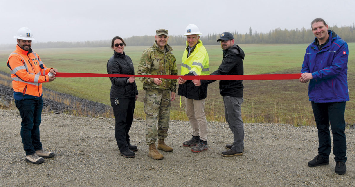 A group of professionals and a military officer in safety gear hold a long red ribbon for an outdoor ceremony in an open field.