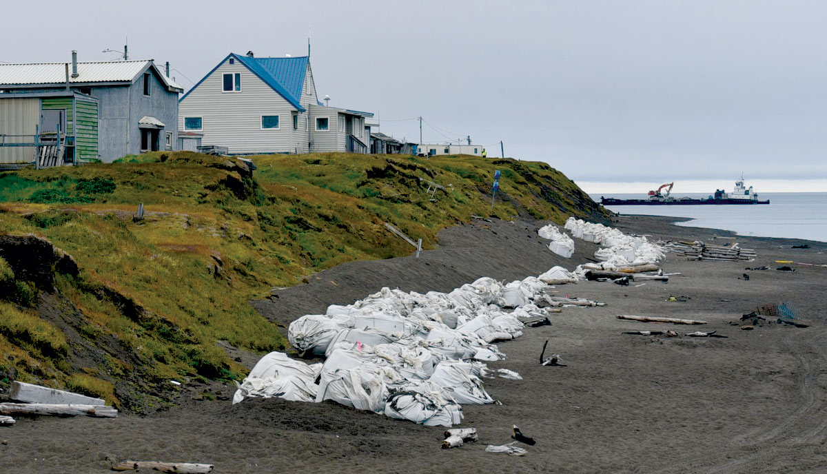 A row of large white sandbags lines a beach to prevent erosion near coastal houses, with a barge visible in the water.