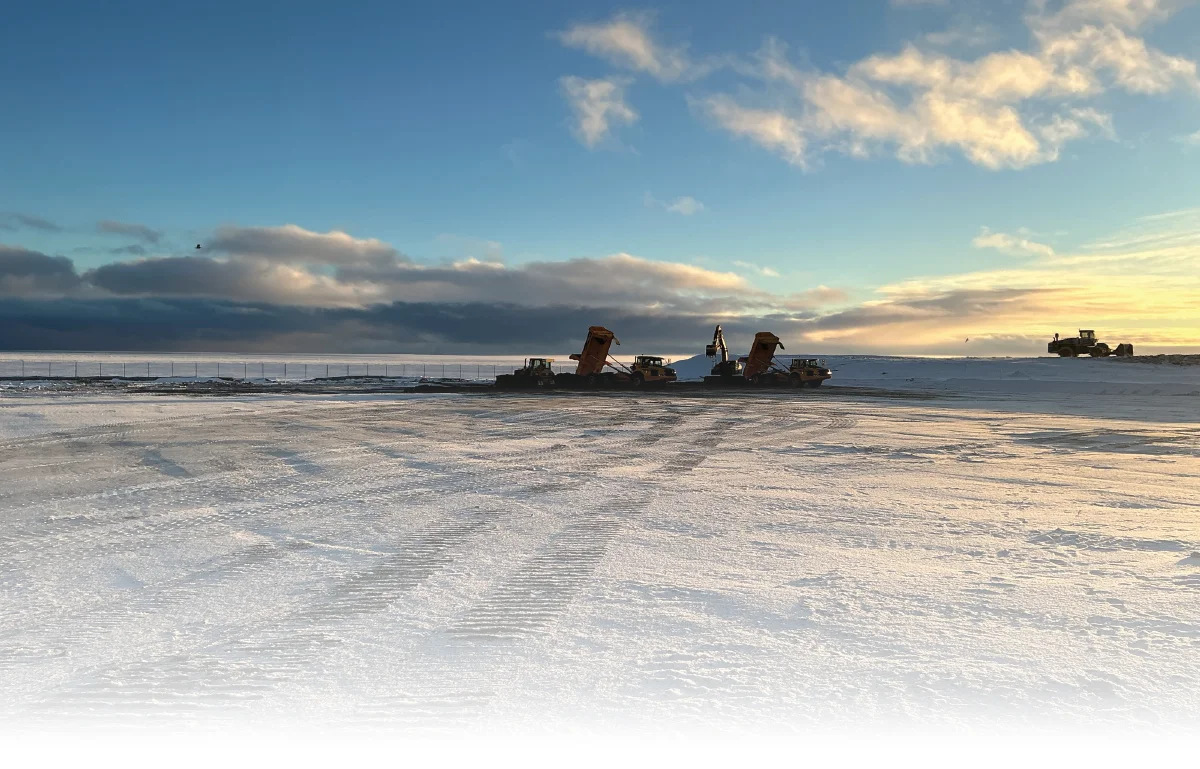 Heavy construction equipment, including dump trucks and an excavator, working on a frozen foundation site in Alaska under a bright Arctic sun
