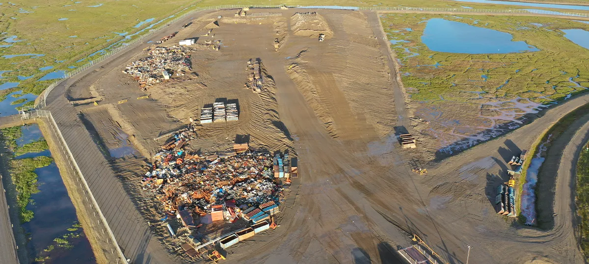 High-angle aerial view of a large industrial construction pad and salvage yard on the Alaskan tundra, featuring organized piles of scrap metal and equipment near a coastal wetland