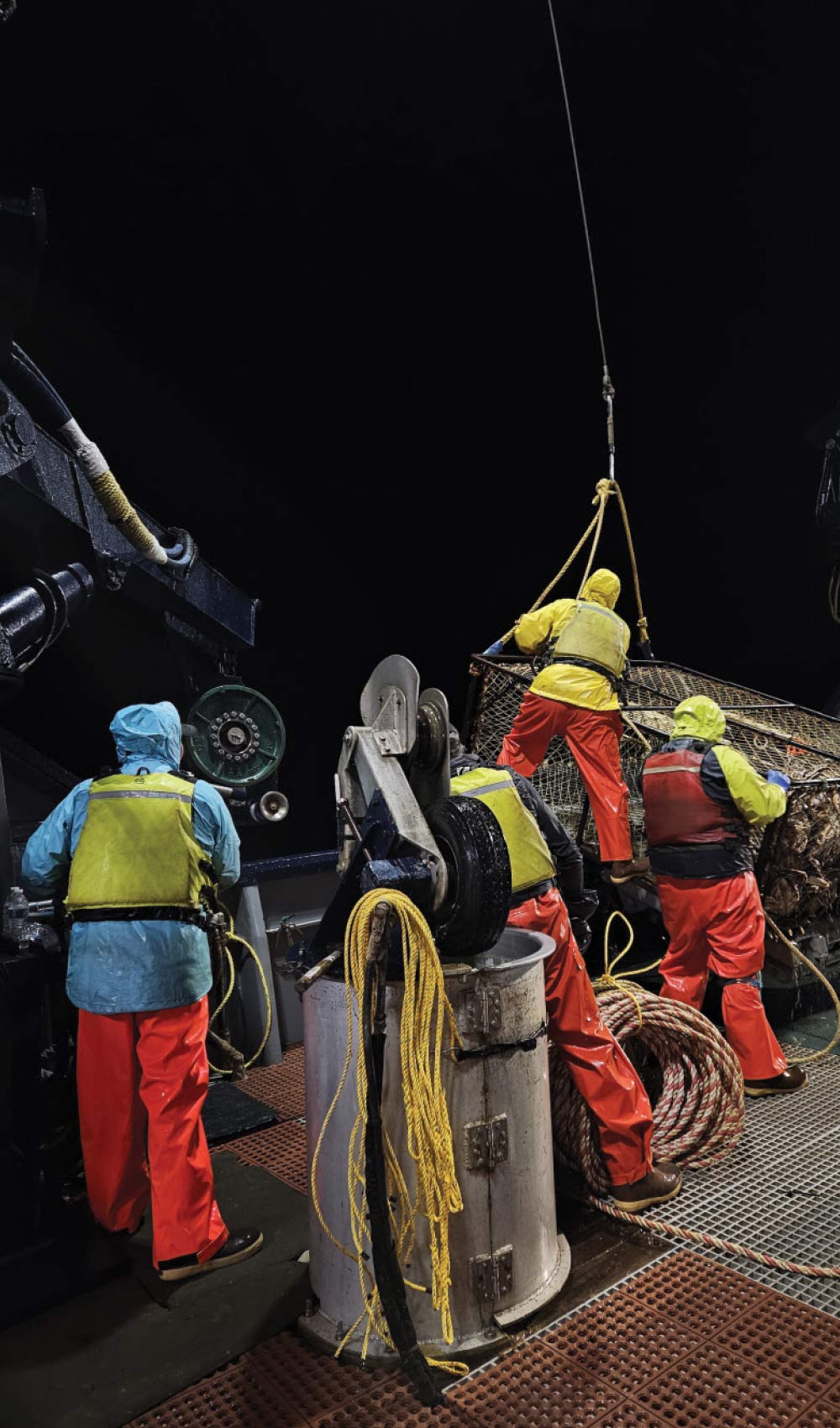 Night view of a commercial fishing crew in yellow and orange waterproof gear working with ropes and a large crab pot on a boat deck.