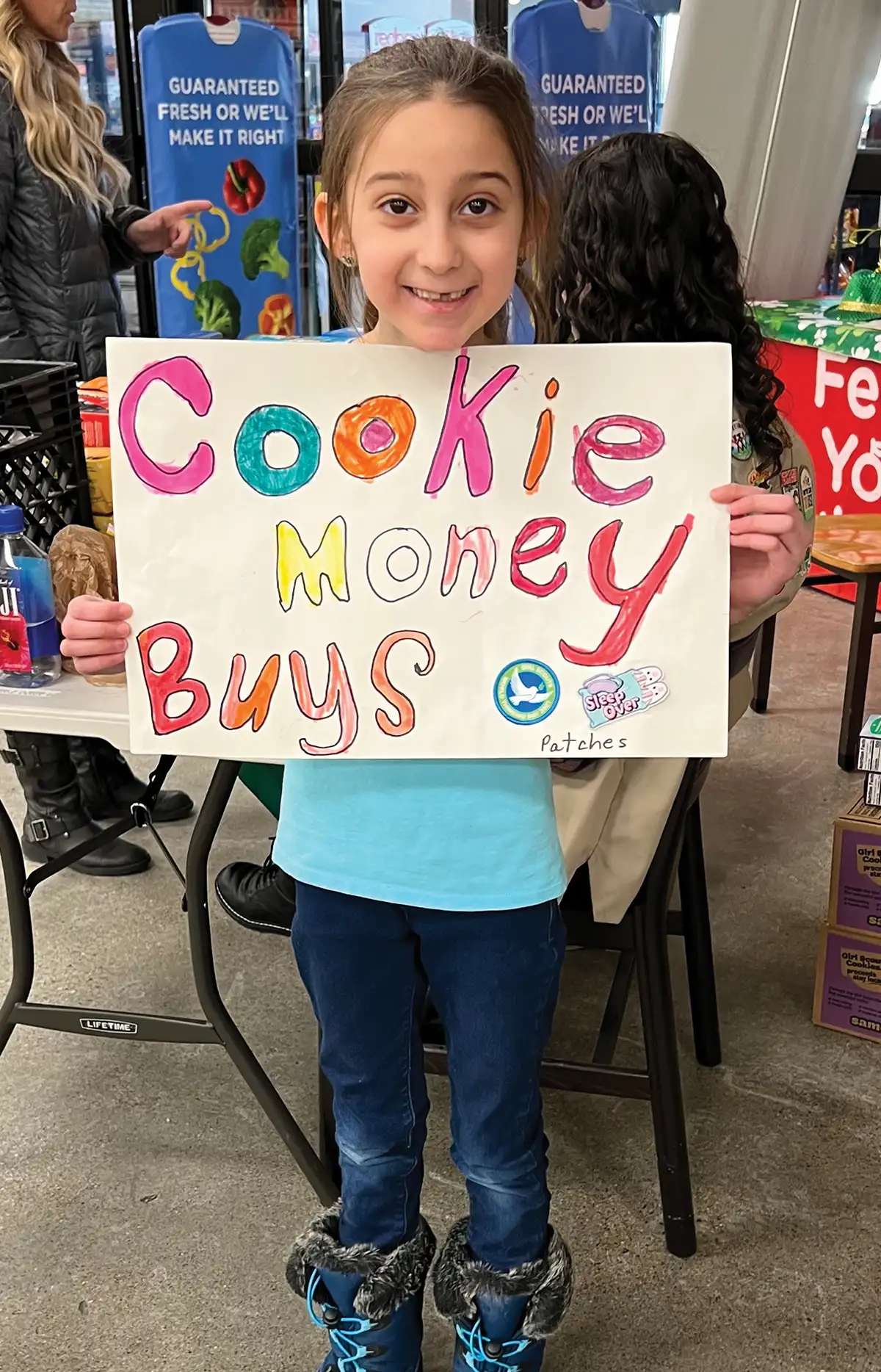 A young Girl Scout smiling while holding a handmade poster that reads "Cookie Money Buys Patches," promoting the troop's fundraising goals during a cookie sale