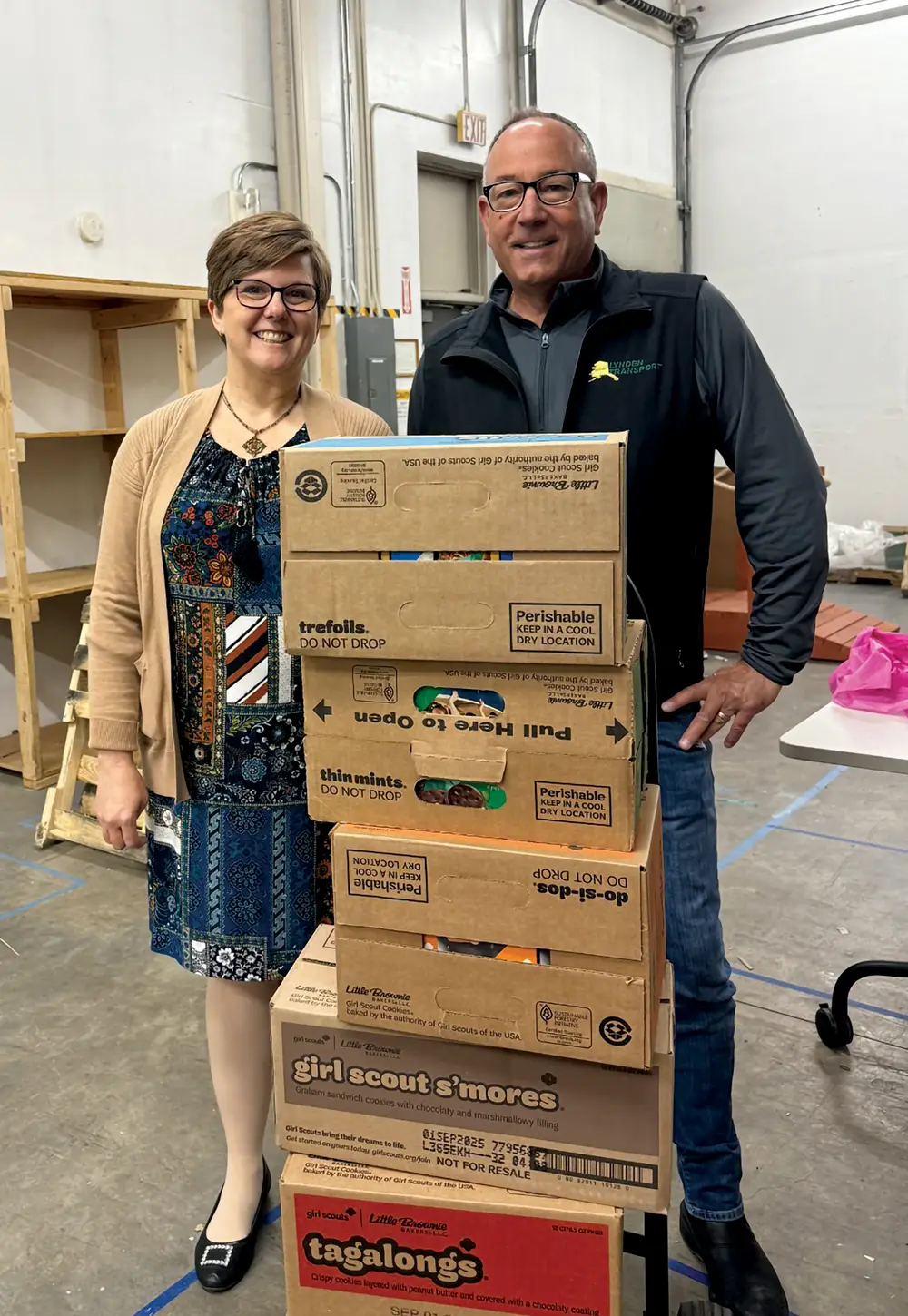 A representative from the Girl Scouts of Farthest North Council and a Lynden Transport official standing with a tall stack of Girl Scout cookie cases, including Thin Mints and Tagalongs, during a distribution event
