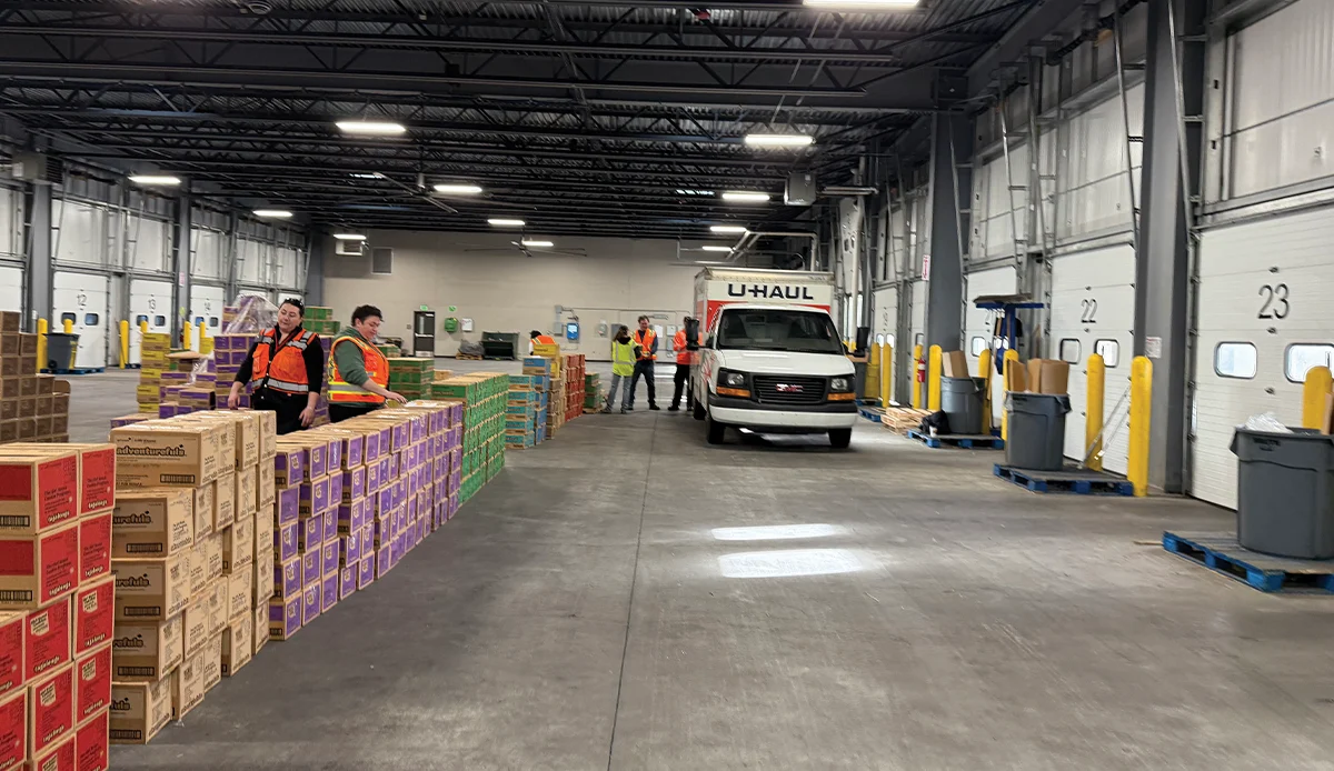 A large industrial distribution warehouse for the Girl Scouts of Farthest North Council, showing long rows of stacked cookie boxes and a U-Haul van parked near loading docks for troop pickup