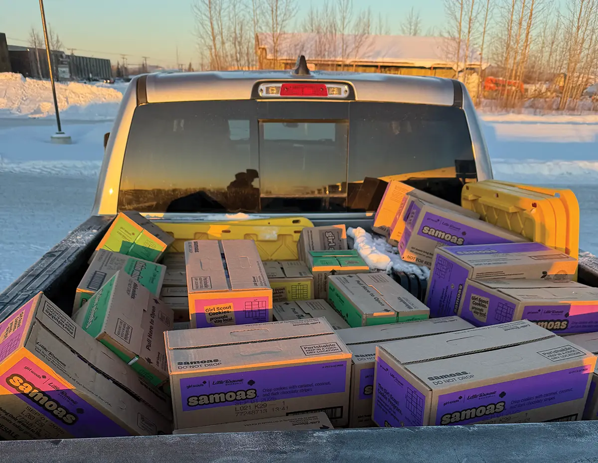 A silver pickup truck bed filled with cases of Girl Scout cookies, including Samoas and Thin Mints, being transported across a snowy Alaskan landscape during the annual cookie distribution