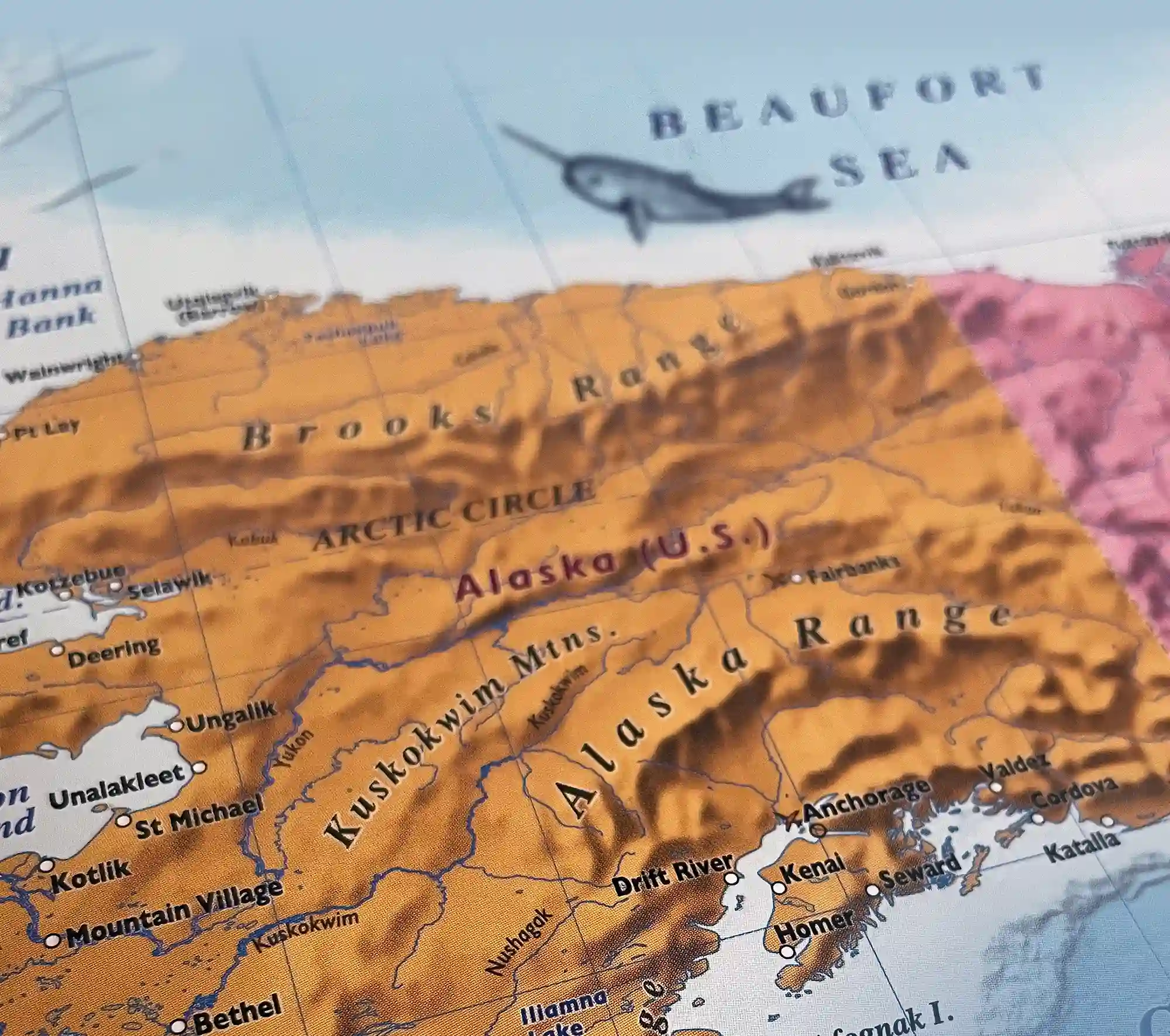 A topographical map of Alaska highlighting major mountain ranges like the Brooks Range and Alaska Range, with a whimsical illustration of a narwhal swimming in the Beaufort Sea