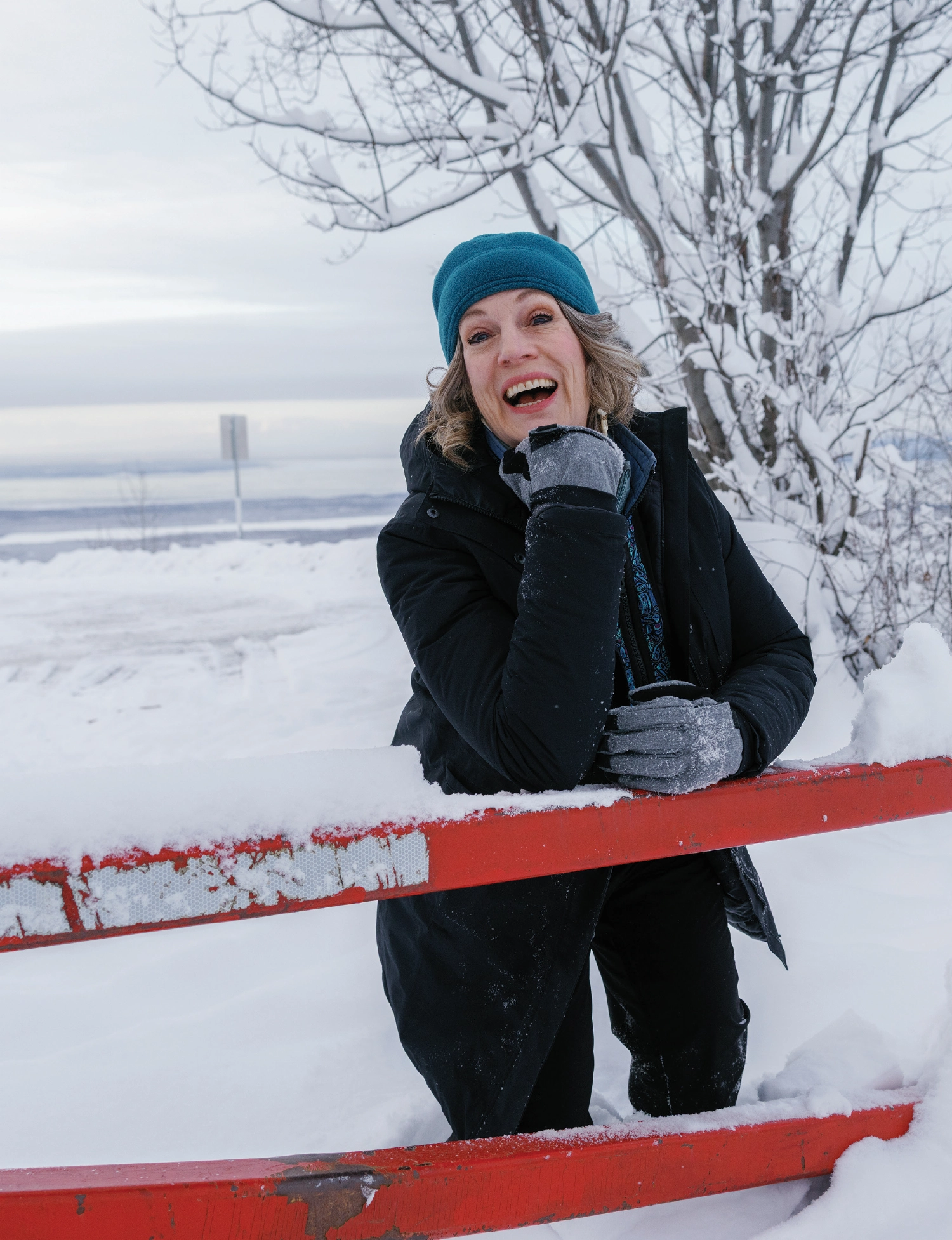 Joelle Hall, a smiling woman elated in joy with her mouth open in a thick black coat and soft teal beanie leaning over a red gate with her right arm placed on top of the red gate while her right hand is leaning on her chin plus her left arm and left hand are resting on top of the red gate as she is standing in a heavy snowy landscape