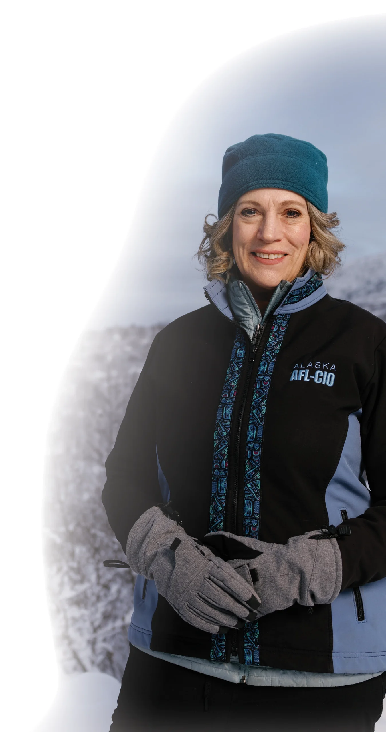 Joelle Hall, a smiling woman in a black and dark muted grey-blue ALASKA AFL-CIO jacket and teal beanie standing in a snowy mountain landscape with her dark grey hand gloves equipped placed over each other