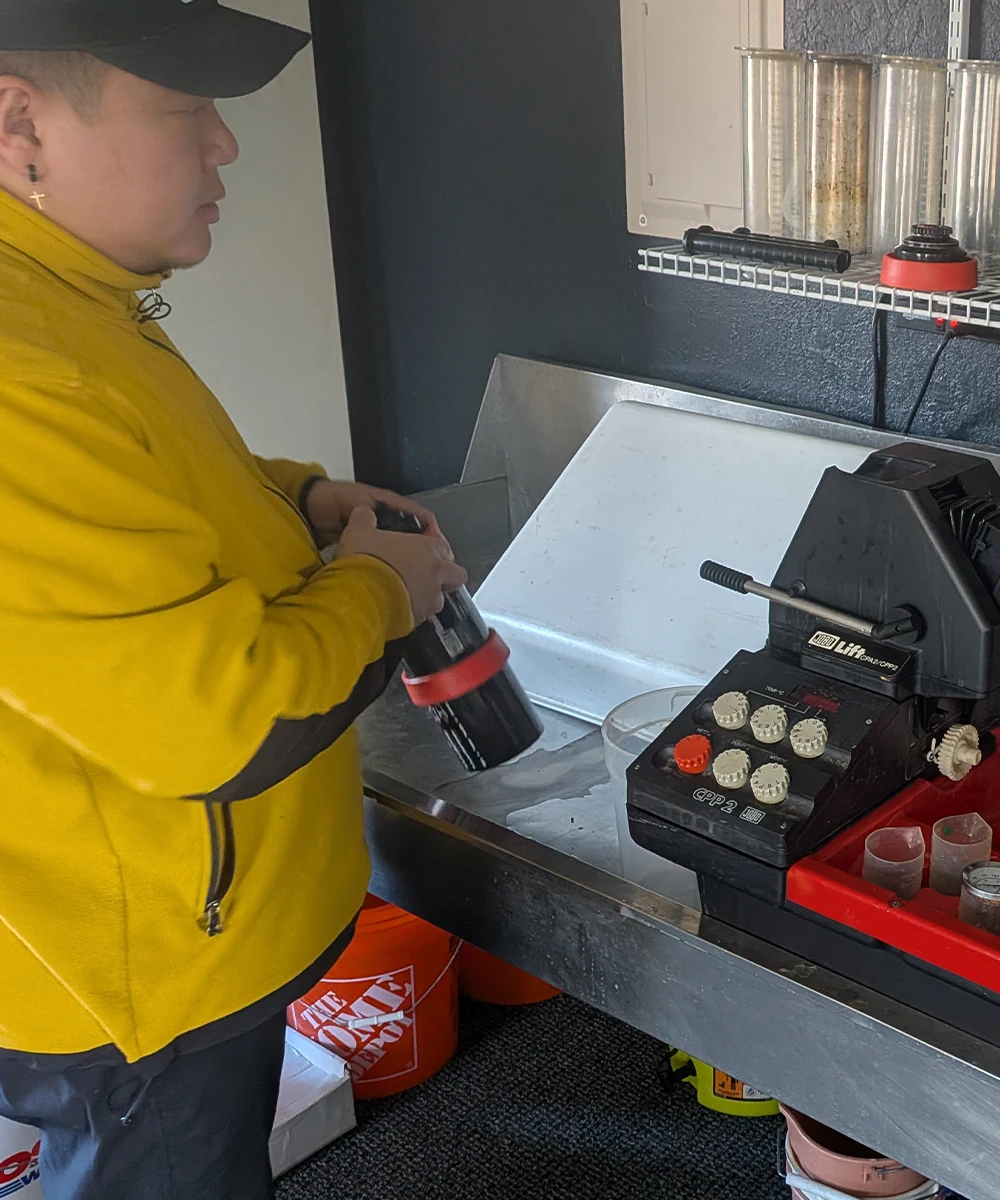 A man in a yellow jacket and black cap holds a black film canister at a stainless steel workstation. The counter features a film processor, and graduated cylinders are visible on a wire shelf above.
