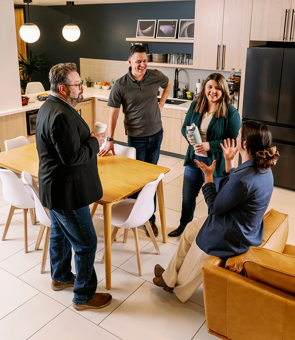 Four colleagues in business-casual attire talk and laugh in a modern office kitchen. One man stands by a light-wood table holding a coffee cup, while another man and woman stand nearby, and a second woman sits on a leather armchair gesturing.