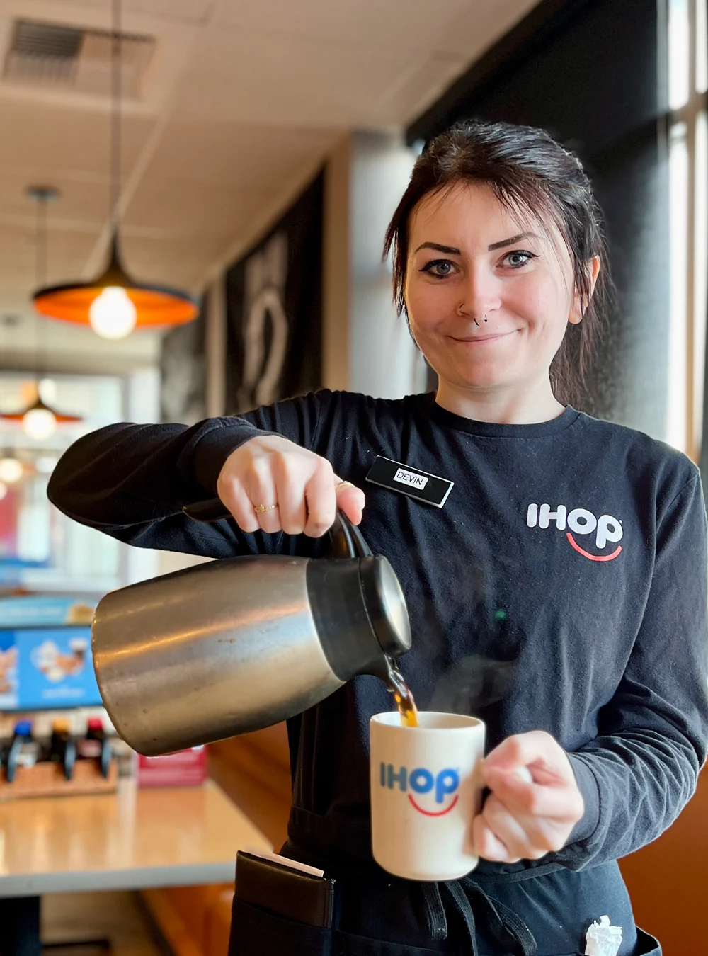 A young IHOP server in a black logo shirt and apron, stands in a restaurant holding a guest check folder. A coffee carafe and mug are on a table in the foreground.