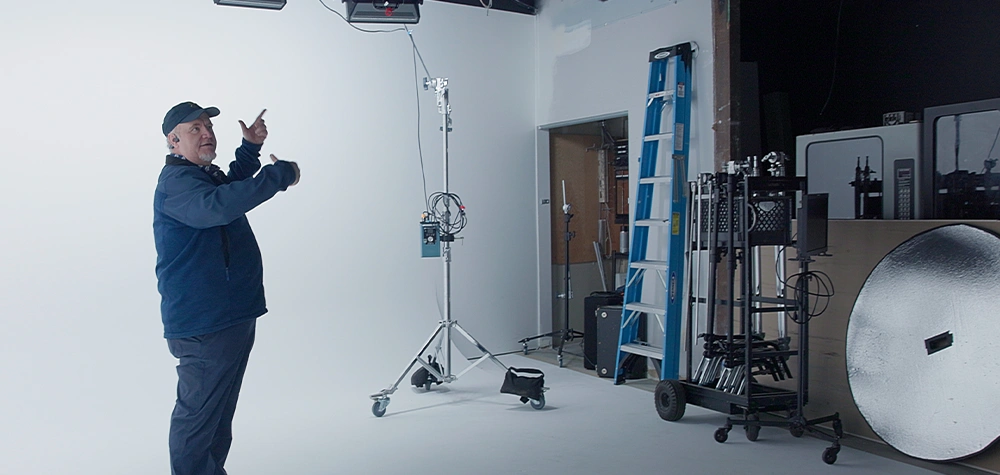 Richard Cooper gestures toward a white cyclorama wall in Frostline Studios. The production space contains a tall light stand on wheels, a blue ladder, equipment carts, and a large circular reflector.