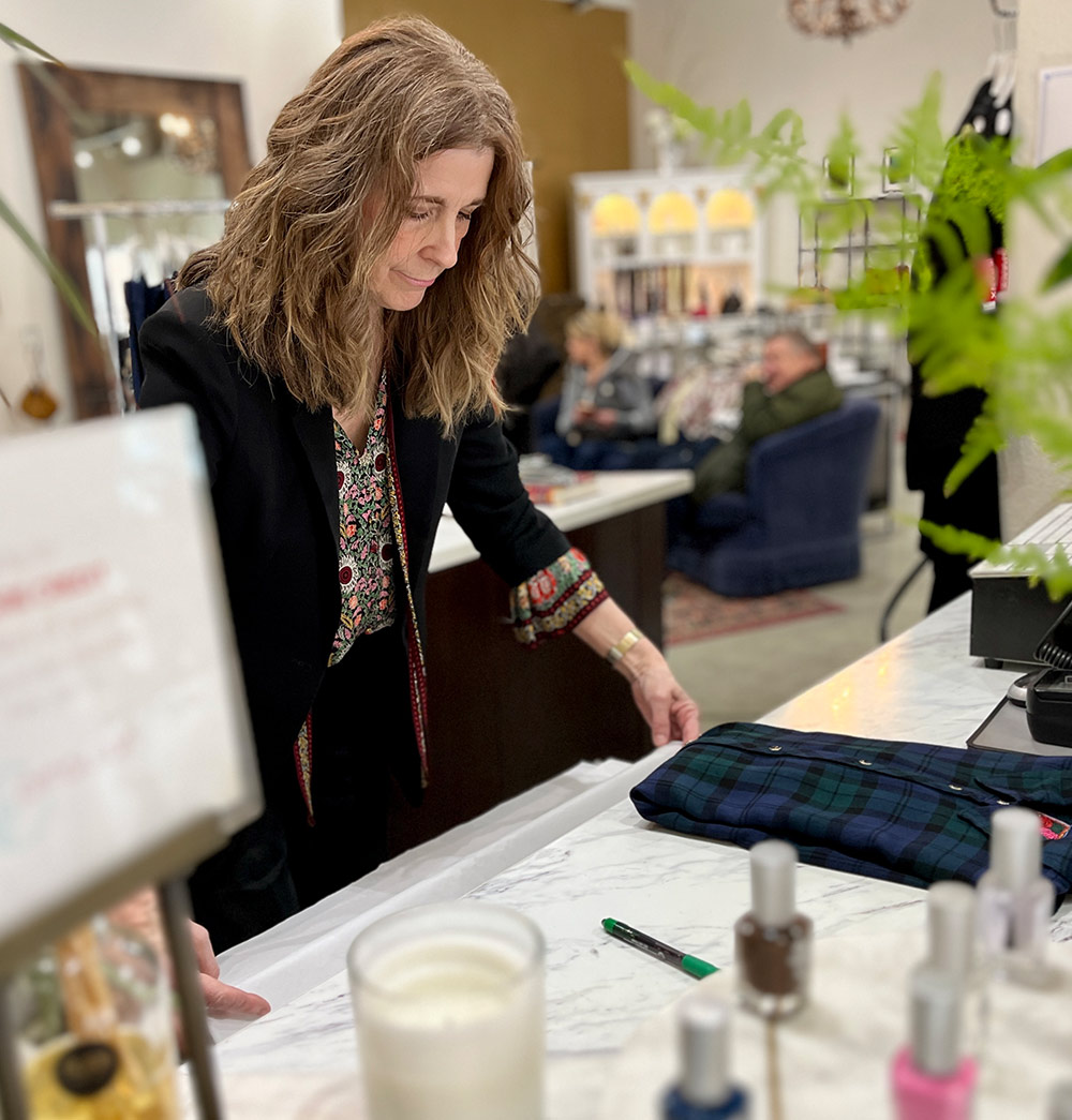 Kim Stalder, wearing a black blazer, folds a blue and green plaid shirt on a marble counter in a boutique. The background shows blurred retail displays and a seated person. Foreground items include a candle and small bottles.