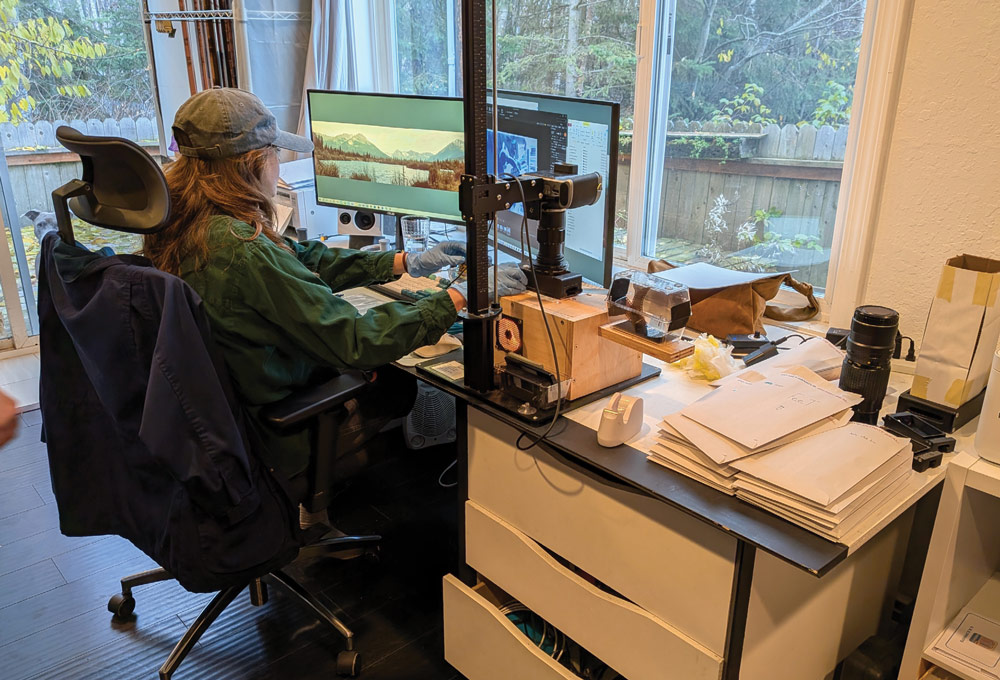 A photographer in a green jacket and cap sits at a desk with dual monitors, using a digital camera mounted on a copy stand to scan and digitize photographic film negatives.