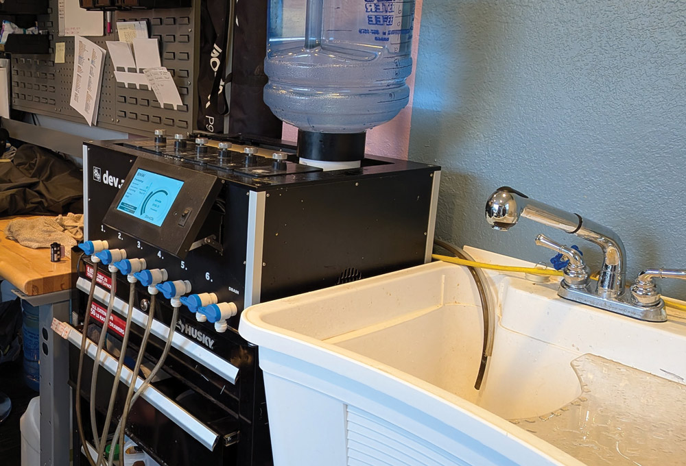 A black automated film processing machine sits on a Husky tool chest next to a white utility sink. A large water jug is mounted on top of the processor for chemical mixing.