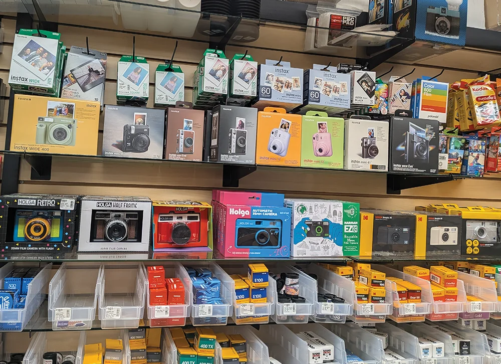 Retail shelves stocked with various Fujifilm Instax cameras, Holga 35mm cameras, and bins of Kodak and Ilford film rolls, showcasing a wide selection of analog photography gear.