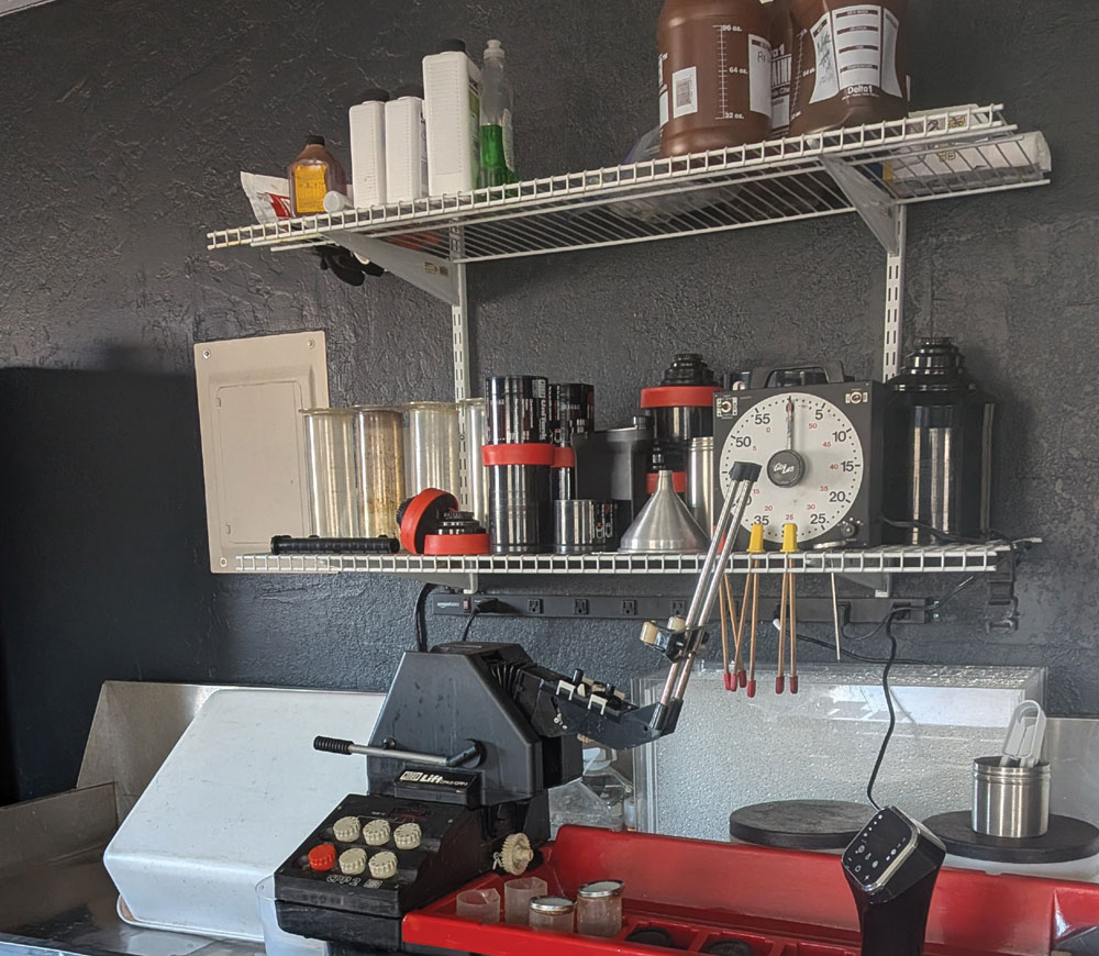 A darkroom workstation featuring a Jobo film processor, a large analog timer, chemical storage bottles on wire shelves, and graduated cylinders used for developing photographic film.