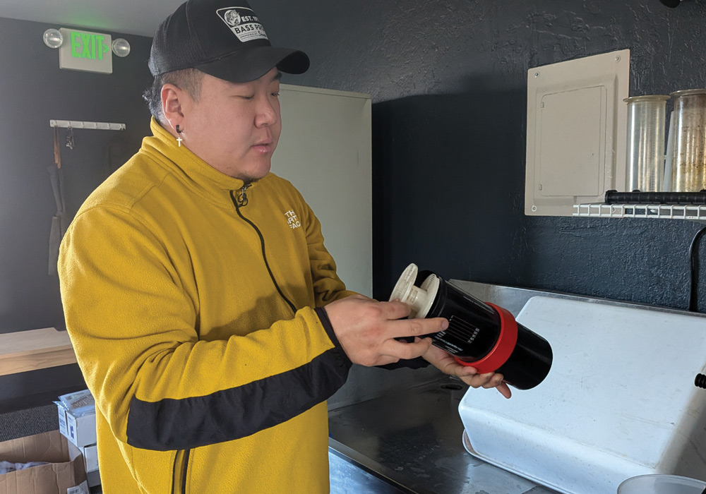 A man in a yellow fleece jacket and black cap stands in a darkroom, carefully loading a roll of photographic film into a black and red plastic developing tank reel.