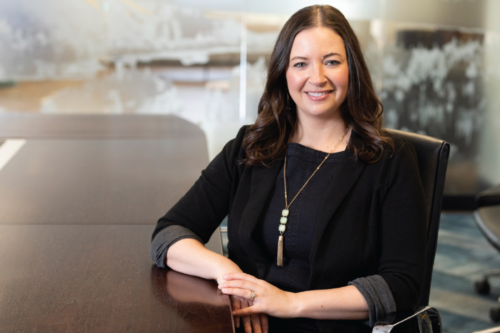 Woman sitting at a conference table, wearing a black blazer and necklace.