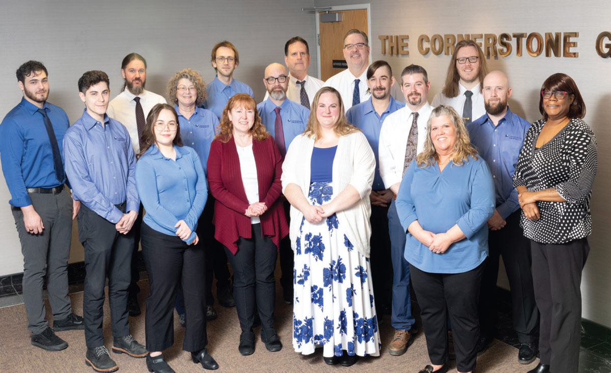 A professional group photo of the Cornerstone Credit Services team, featuring sixteen employees smiling and standing in an office hallway under a sign that partially reads "THE CORNERSTONE".