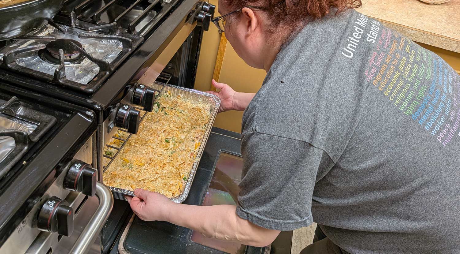 A person in a grey t-shirt slides a large, foil-covered catering tray into a commercial-grade oven.