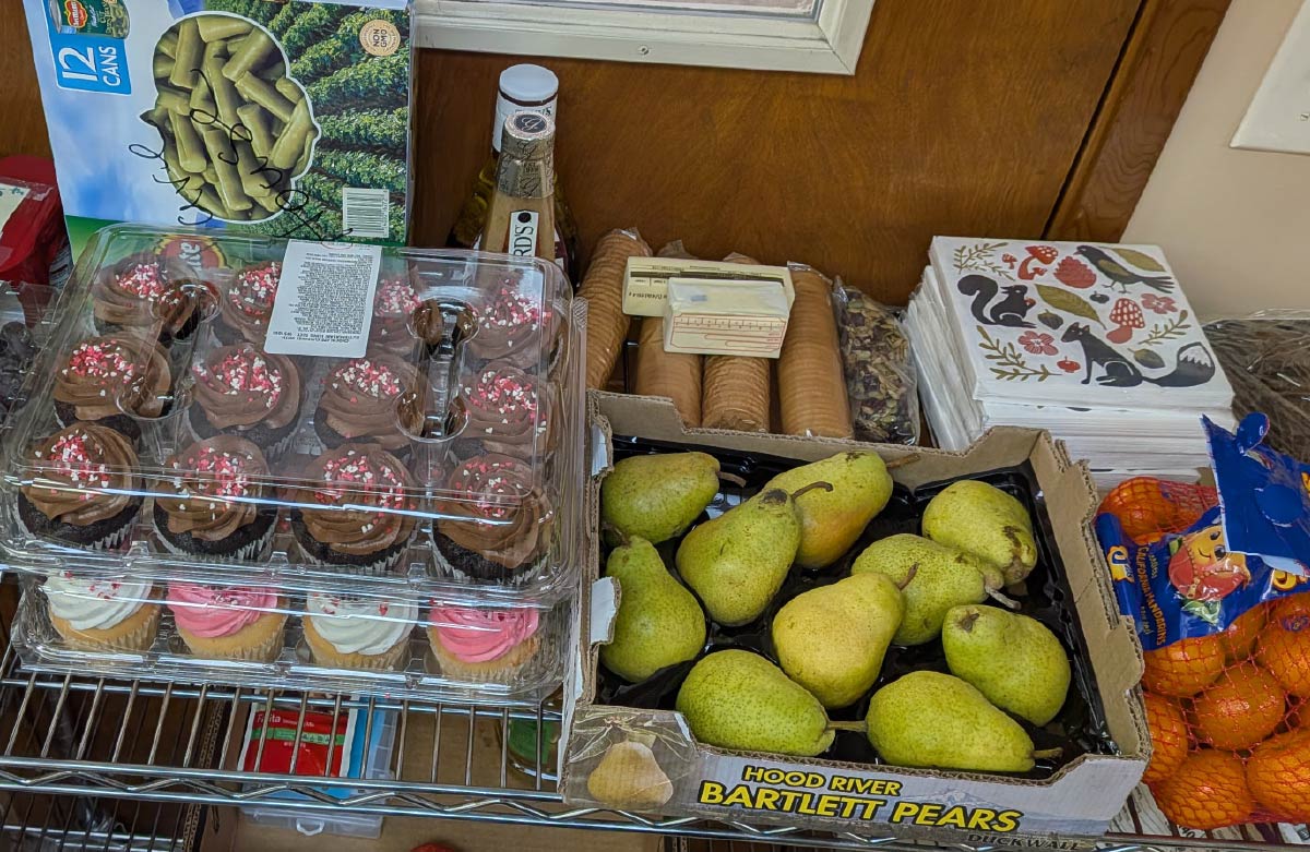 A tabletop display of donated food including fresh Bartlett pears, chocolate cupcakes, oranges, and crackers.