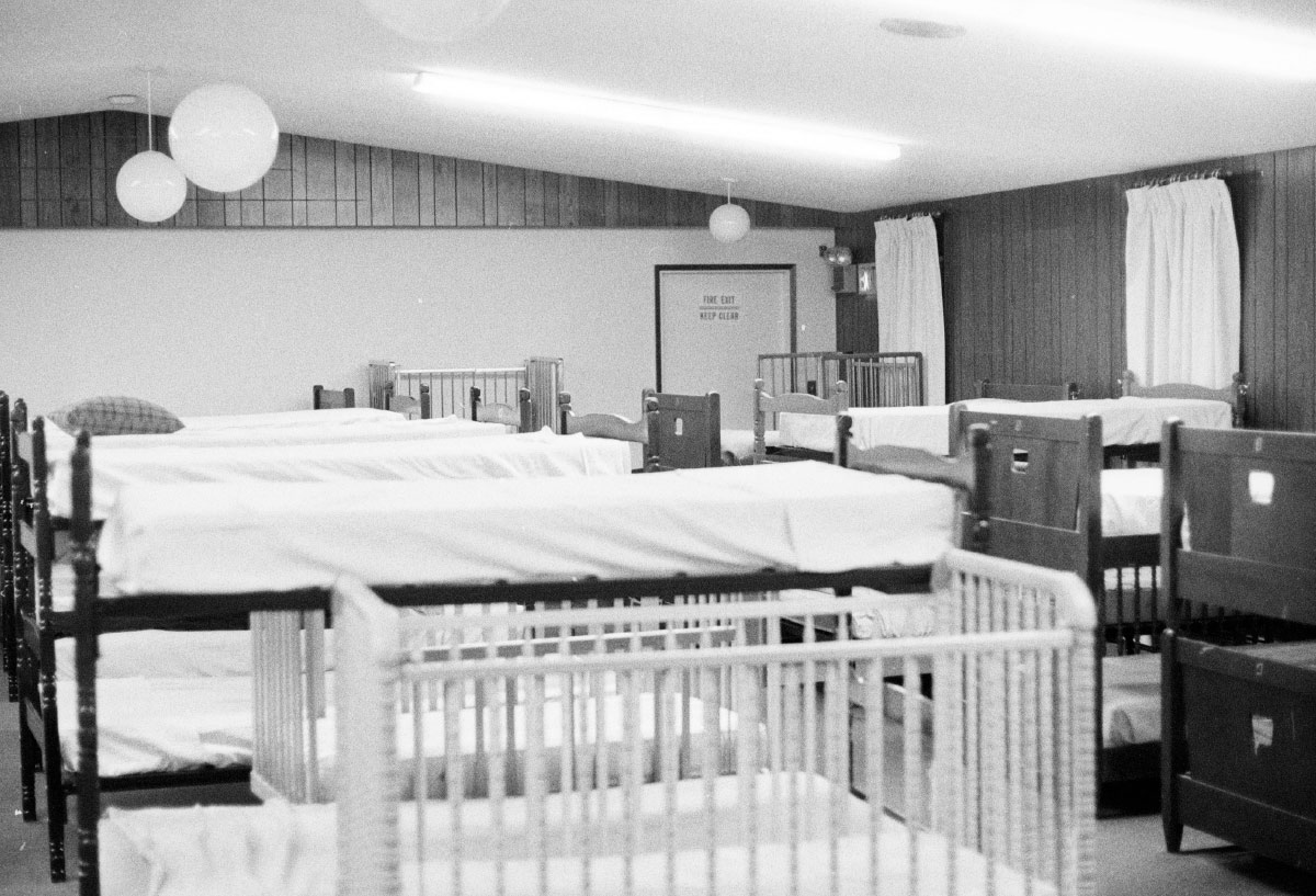 A black-and-white photo showing an indoor communal sleeping area filled with several wooden cribs and cots.