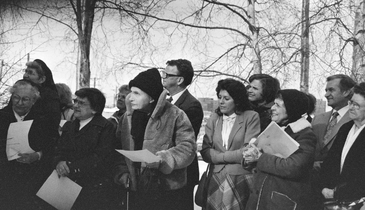 A nostalgic black-and-white group portrait of people standing outdoors, many holding papers, with snow and bare trees in the background.