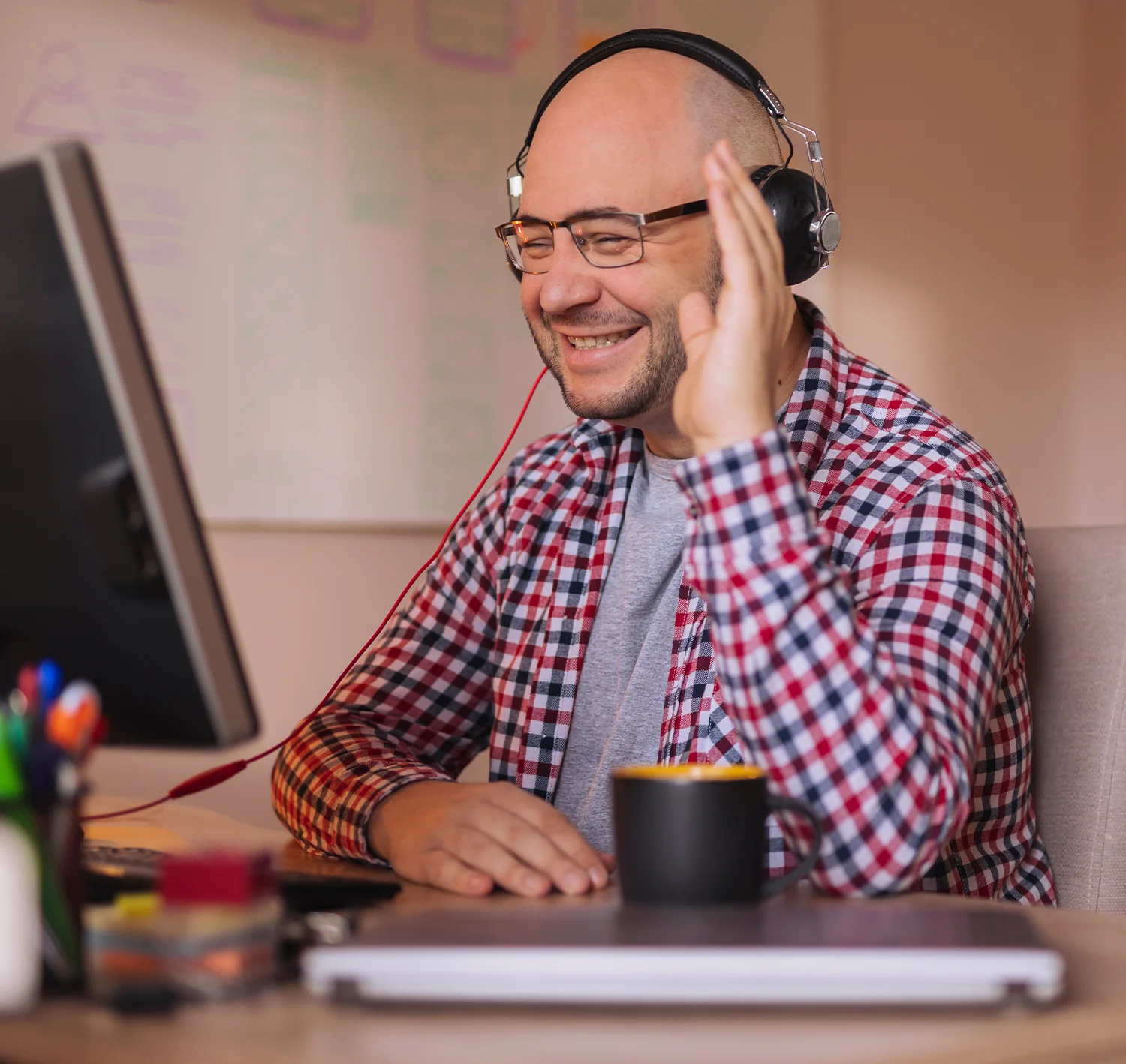 A smiling man wearing headphones and a plaid shirt waves at his computer screen while sitting at a desk with a coffee mug