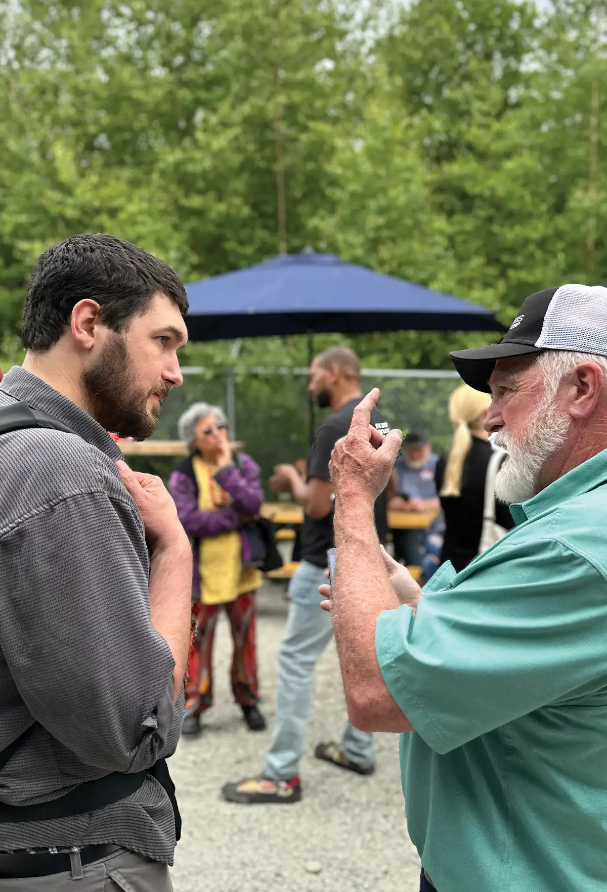Two men in a focused conversation at an outdoor community event with a blurred background of people and trees.