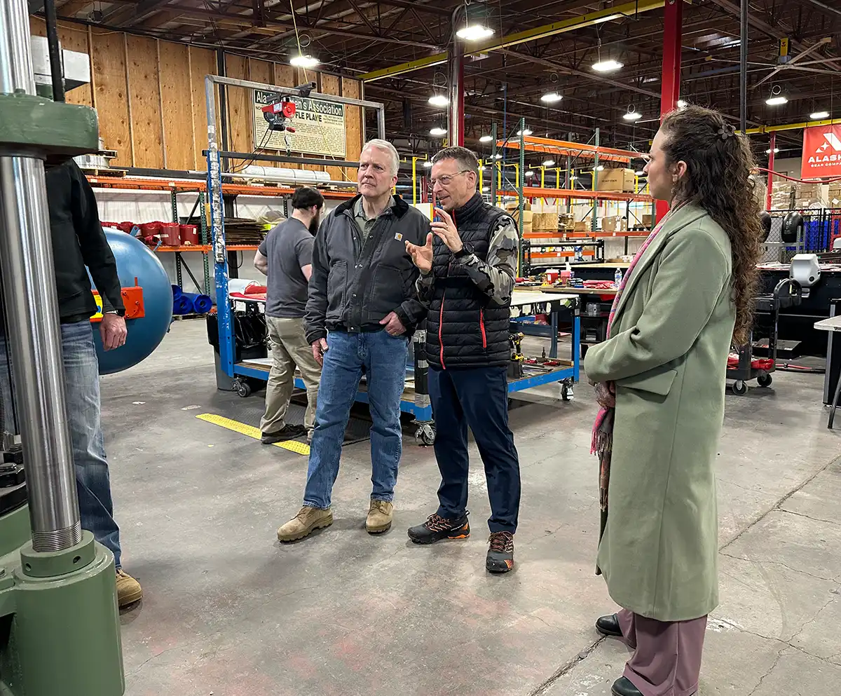 A group of four people in a large industrial warehouse discussing machinery near storage racks and a blue workbench