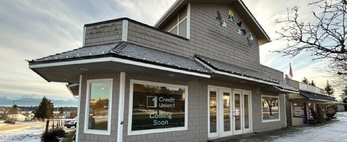 A grey shingled building with a sign in the front window that reads "Credit Union 1 Coming Soon" set against a snowy mountain backdrop.