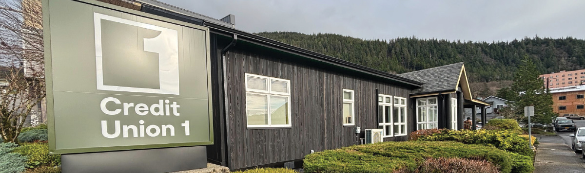 An outdoor monument sign for Credit Union 1 stands in front of a dark, rustic wooden office building surrounded by greenery and a forest.